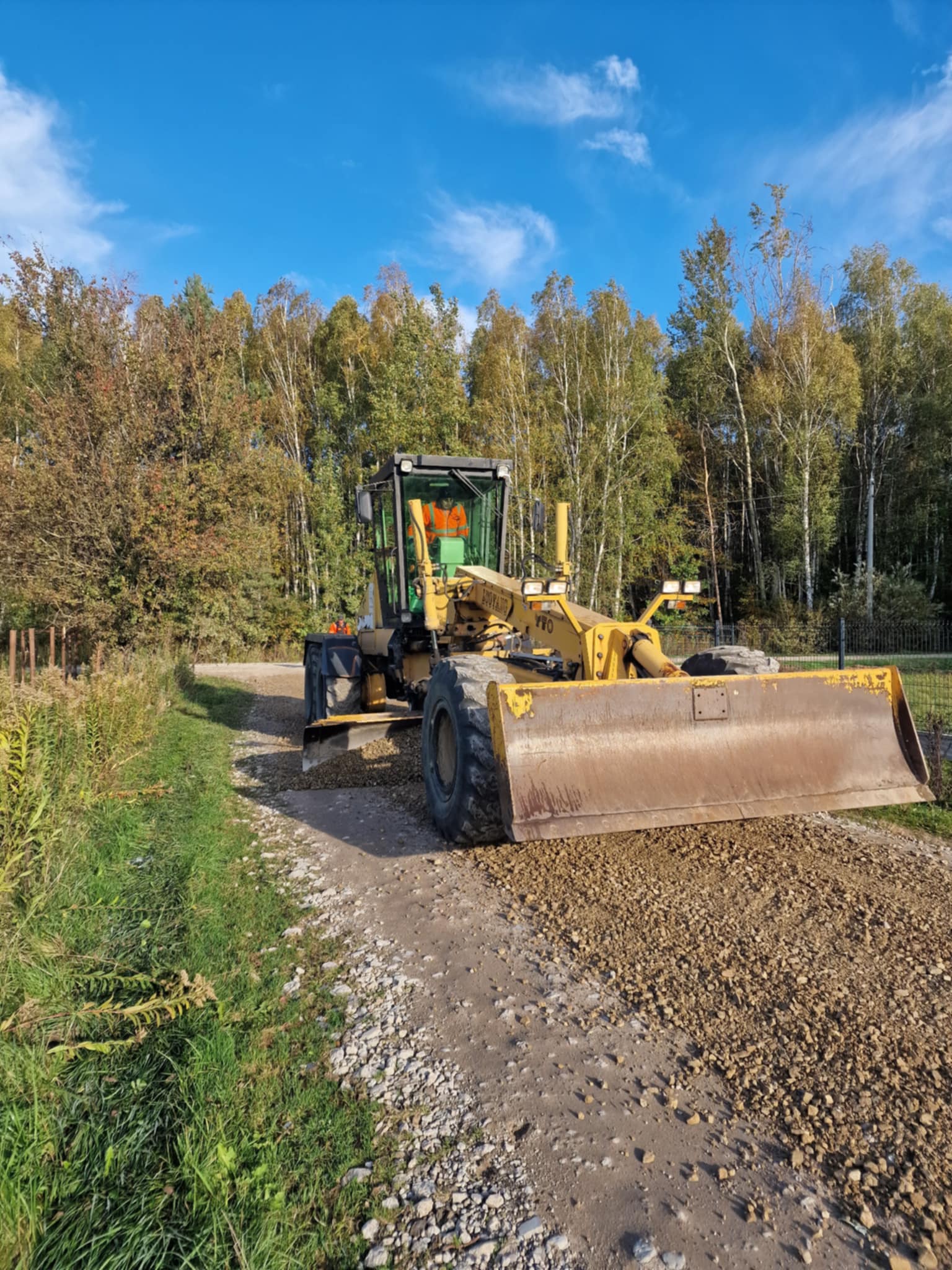 Na zdjęciu widoczna jest żółta równiarka. W niej znajduje się operator. W tle zdjęcia drzewa - brzozy. Po lewej stronie zdjęcia pobocze porośnięte trawą.