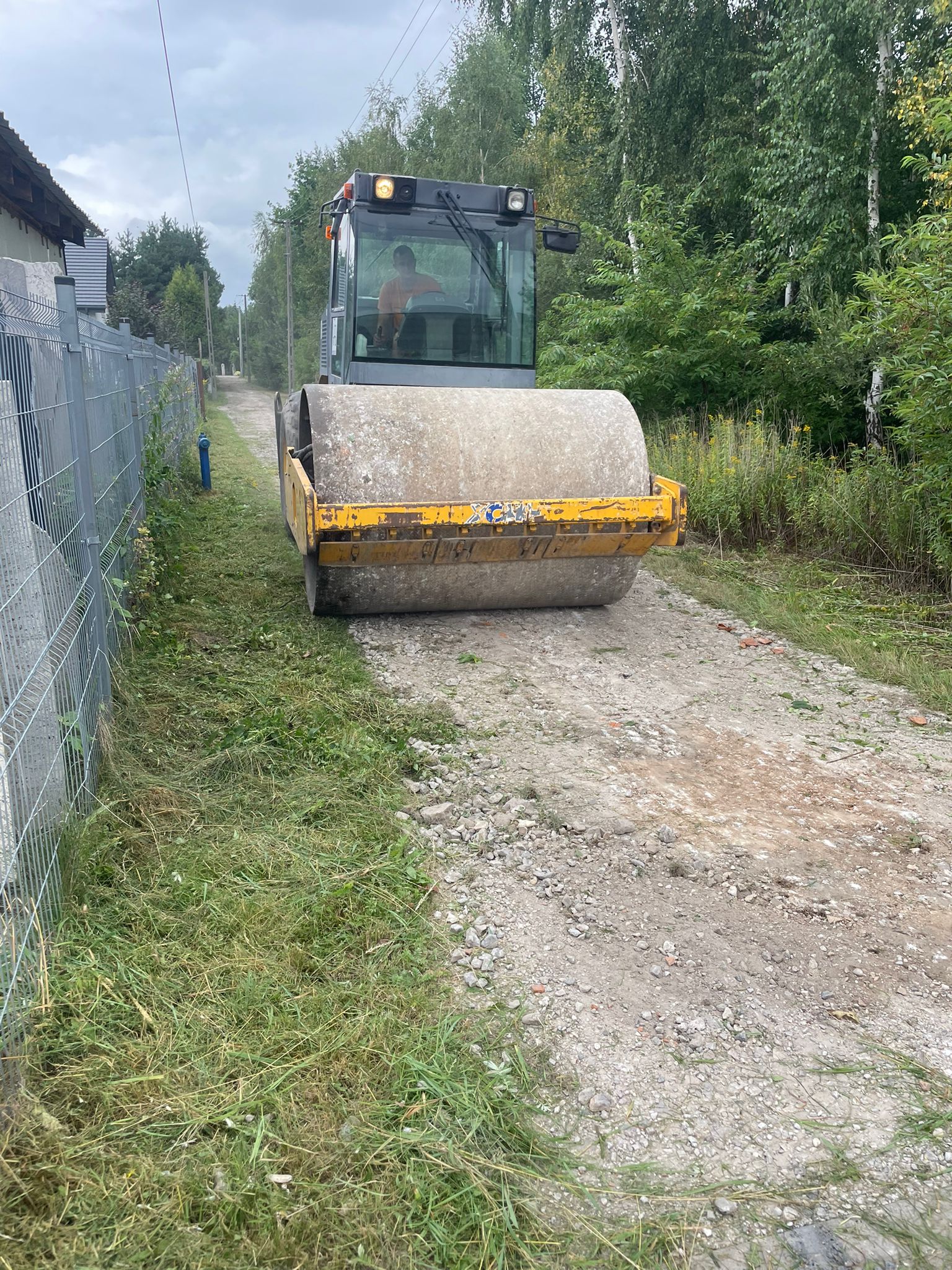 Na pierwszym planie zdjęcia znajduje się walec równający drogę. W tle po prawej stronie znajdują się duże drzewa (sosny), po lewej stronie metalowe ogrodzenie.