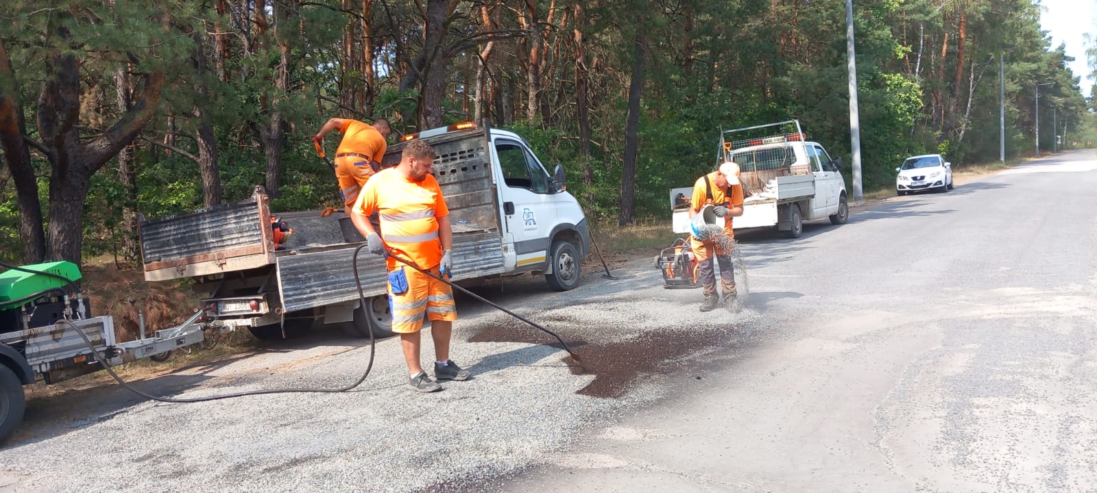 Na pierwszym planie znajduje się pracownik Spółki rozgrzewający wysypany na drodze grys. Z prawej strony zdjęcia także znajduje się pracownik Spółki, który trzyma w rekach łopatę. Pracownik stoi na przyczepie auta, jest odwrócony tyłem i pochylony do przodu. Z lewej strony stoją dwa białe auta oraz pracownik Spółki wysypujący grys na drogę.