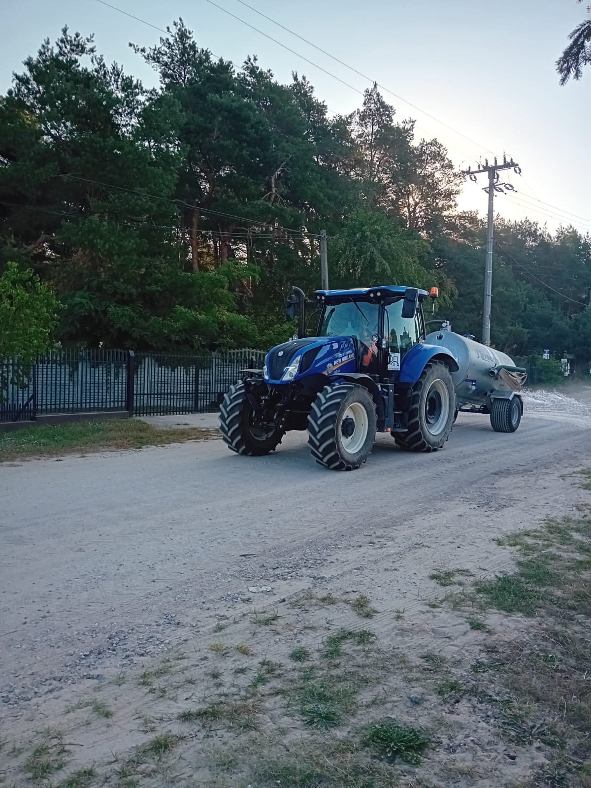 Na pierwszym planie znajduje się niebieski traktor marki New Holland wraz z beczkowozem jadący po gruntowej drodze. Po lewej stronie widać metalowe grodzenie, po prawej pobocze porośnięte trawą.