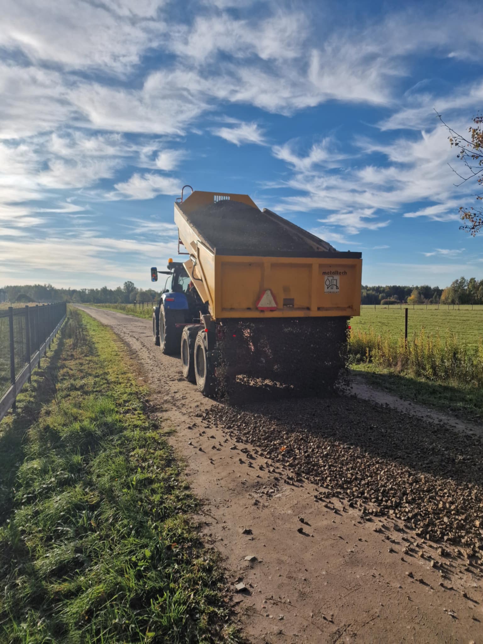 Na pierwszym planie znajduje się odwrócony tyłem traktor z naczepą. Z naczepy wysypuje się kruszywo, jest uniesiona.Po prawej stronie zdjęcia znajdują się pola, po lewej widoczne jest ogrodzenie.