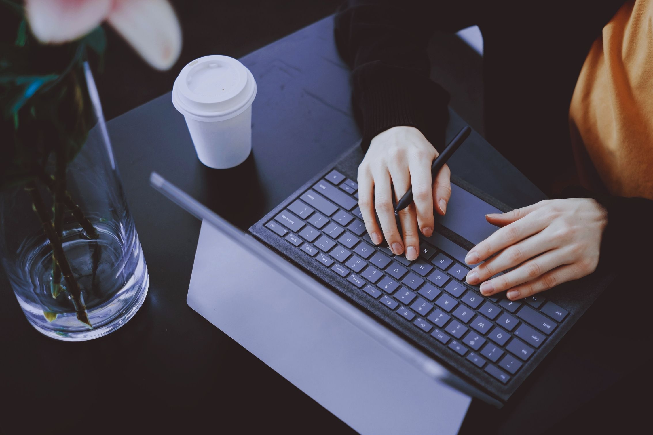 Po prawej stronie widoczne są dłonie kobiety, która pisze na laptopie. Po lewej stoi wazon z kwiatami i biały kubek.