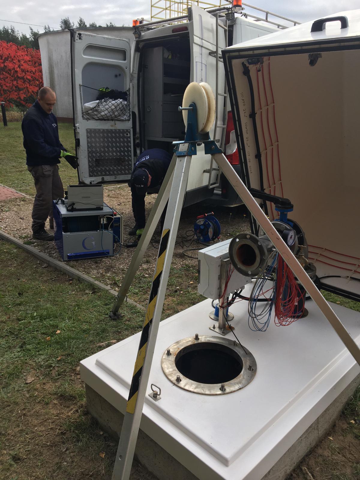 Po lewej stronie kuca mężczyzna, który ma zwrócony wzrok w stronę komputera stojącego na metalowej skrzynce, w tle tył samochodu oraz dwie inne postacie. Po prawej otwarta studnia głębinowa nad którą stoi statyw do kamery.